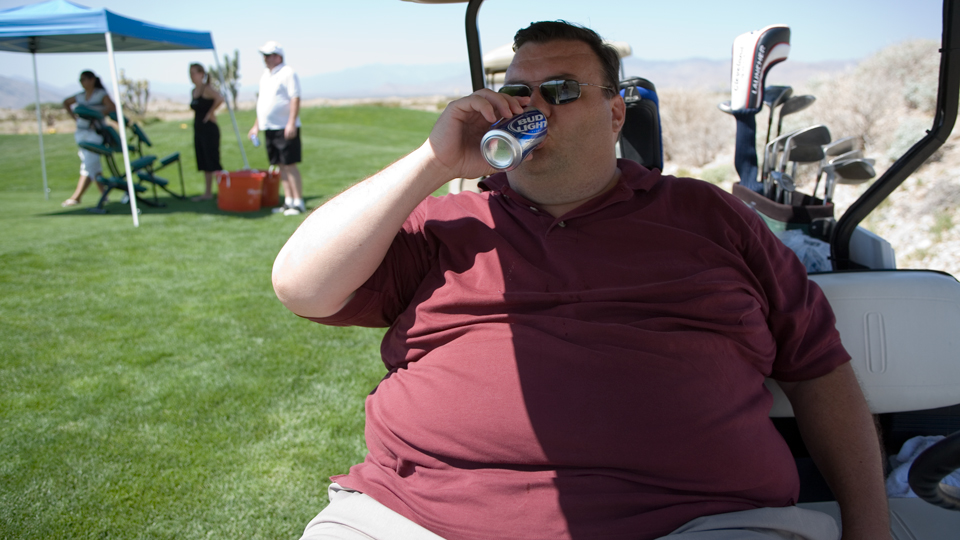 Obese golfer sitting on a golf cart drinking Bud Light. 