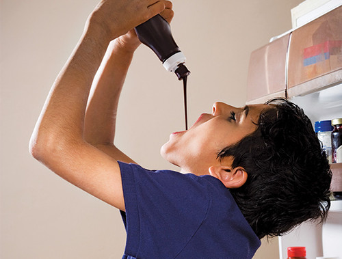 Preteen standing in front of an open refrigerator raising an upturned Hershey's chocolate squeeze bottle over his mouth with both hands. Liquid chocolate is pouring into his mouth. [Formatted]